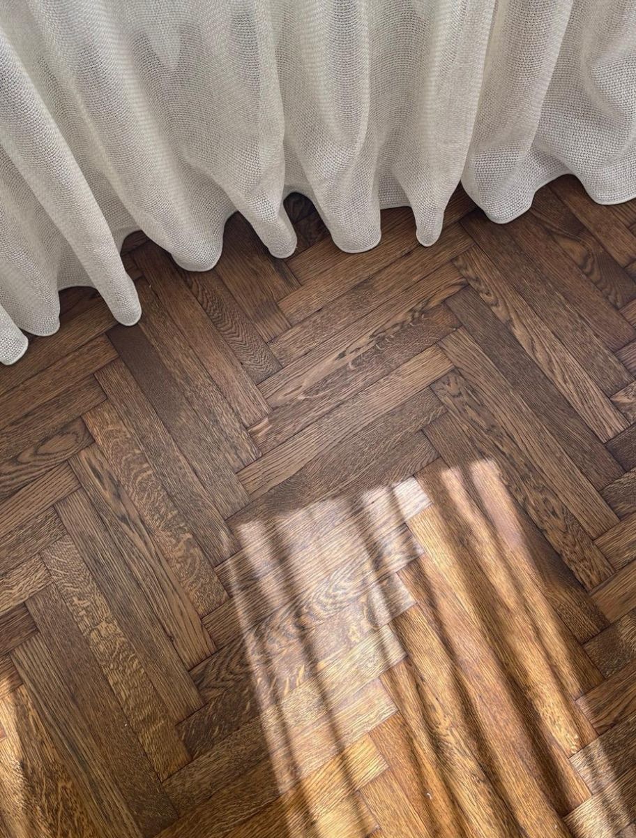 Sunlight casting shadows on a herringbone-patterned wooden floor treated with Paint Eco Tinted Hard Wax Oil, complemented by a white curtain.