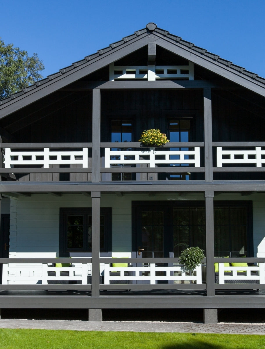 Modern wooden house exterior with a dark-painted facade and white railings, coated with linseed oil paint. Spacious veranda with green accents and flower pots.