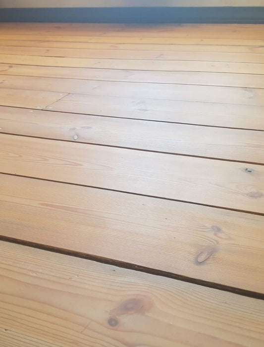 A close-up view of a wooden floor with a natural, light finish, showcasing visible wood grain and knots. The surface appears smooth and well-treated, possibly with a protective wood oil or wax.