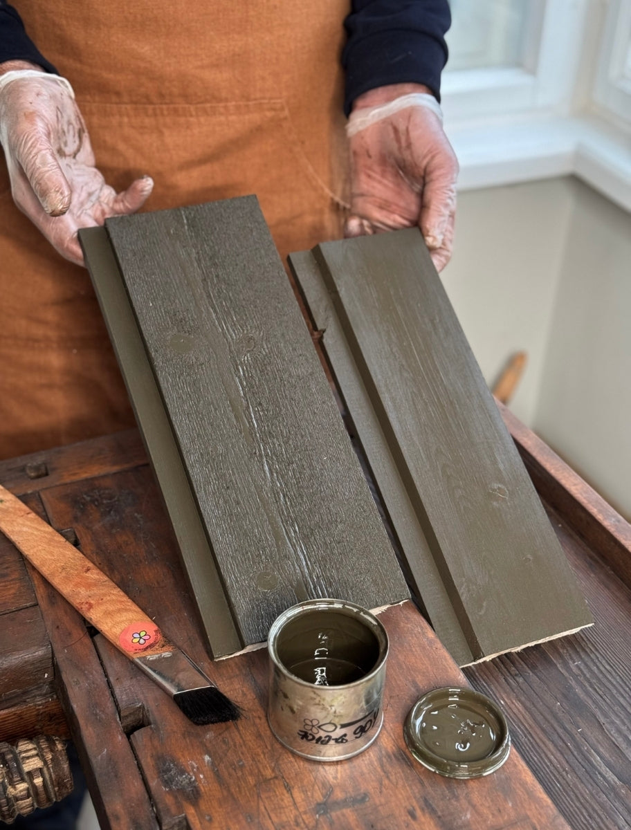 A person holding wooden boards painted with Paint Eco natural linseed oil paint in Dark khaki shade, with an open can beside them.