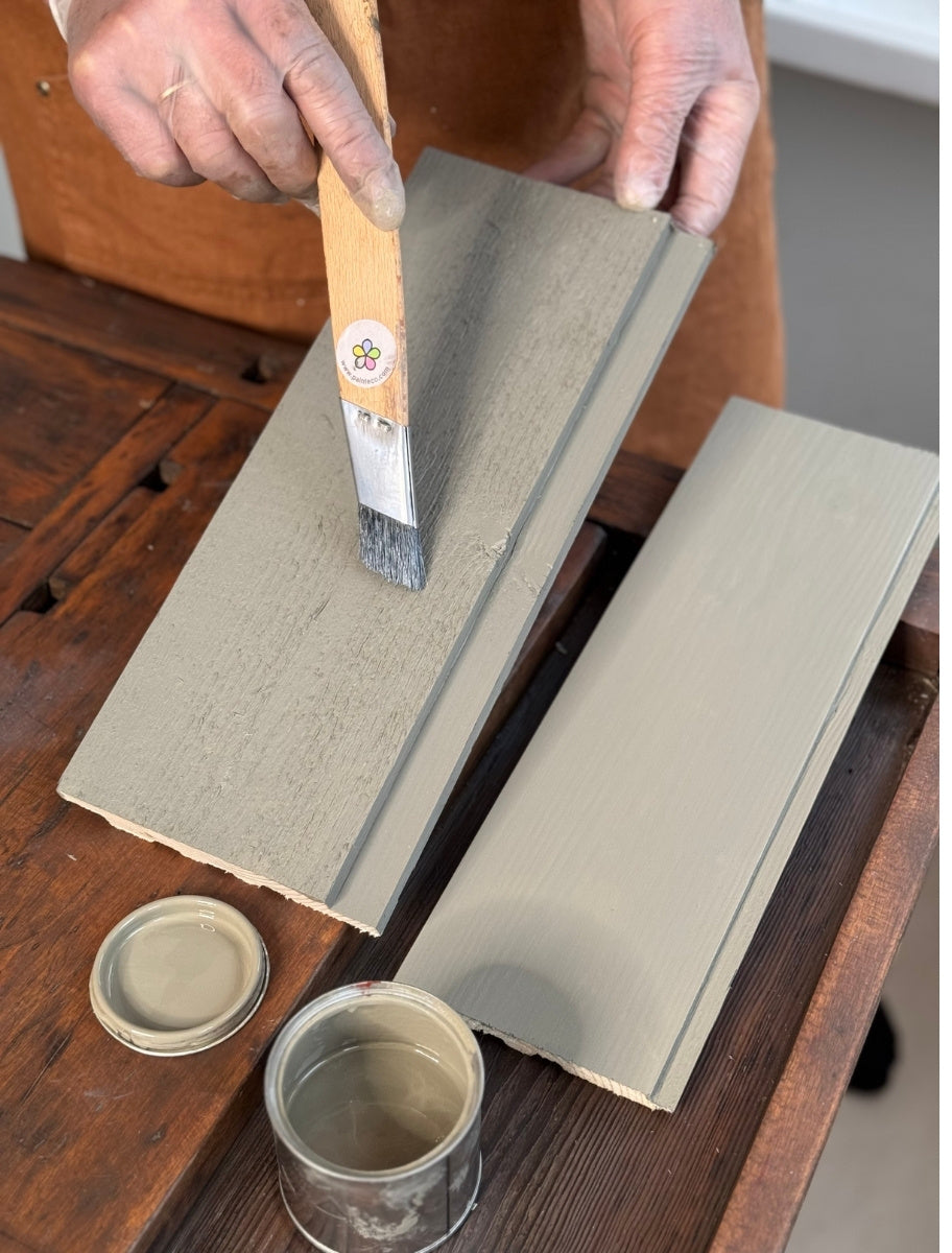 A person using a paintbrush to apply Paint Eco natural paint Pigeon shade on a wooden board, with an open can beside them.